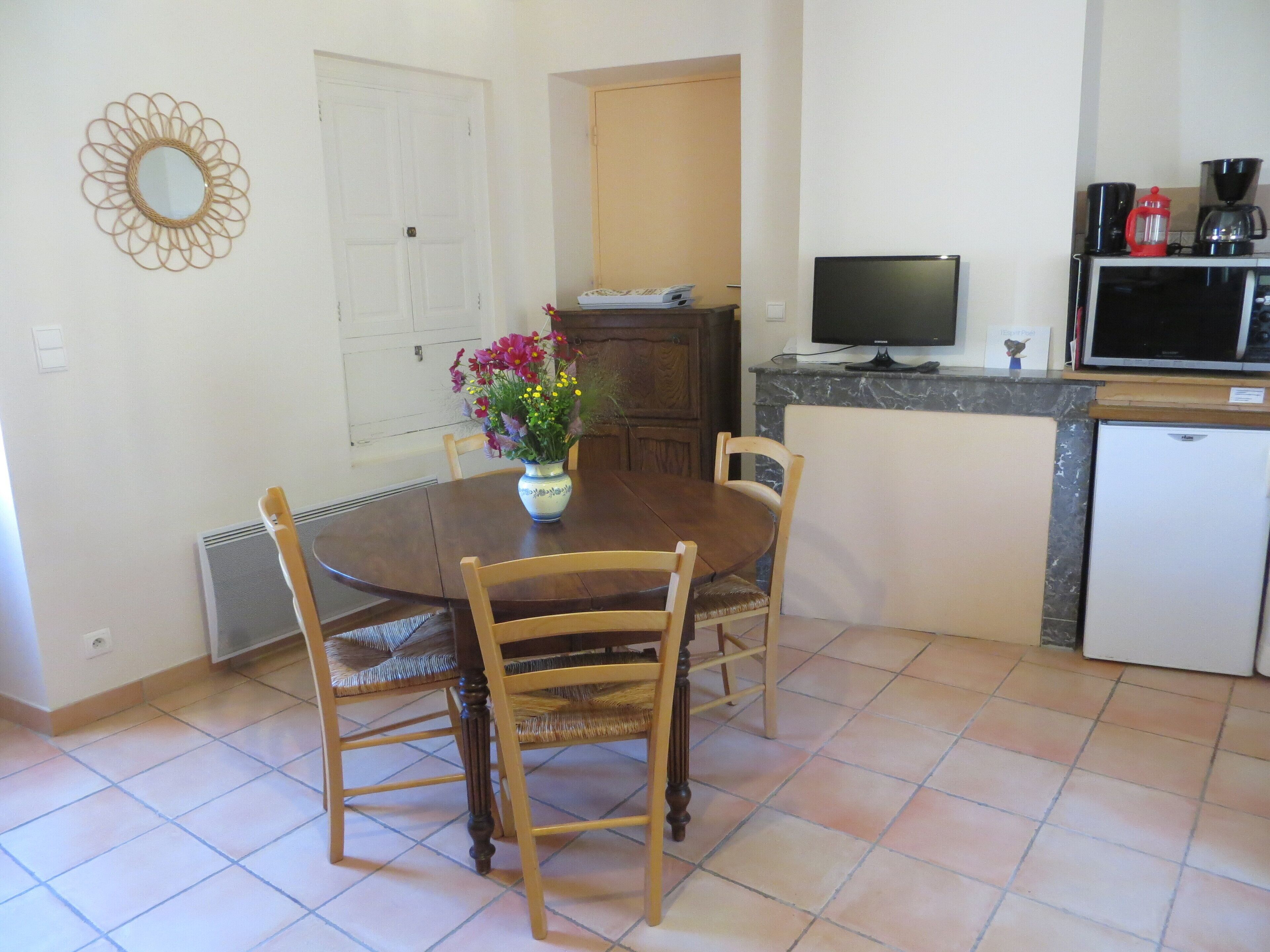 Uma sala de jantar com uma mesa redonda, quatro cadeiras, um vaso com flores, uma televisão e uma cafeteira.