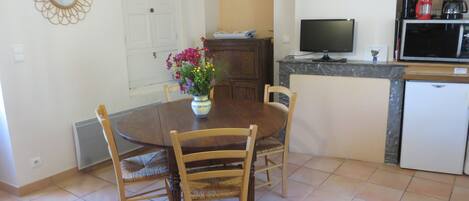 Uma sala de jantar com uma mesa redonda, quatro cadeiras, um vaso com flores, uma televisão e uma cafeteira.
