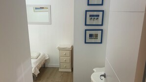 Ein schmaler Flur führt zu einem Schlafzimmer mit einem Bett, einem Nachttisch und einer kleinen Kommode. An der Wand über der Kommode hängen drei gerahmte Bilder.