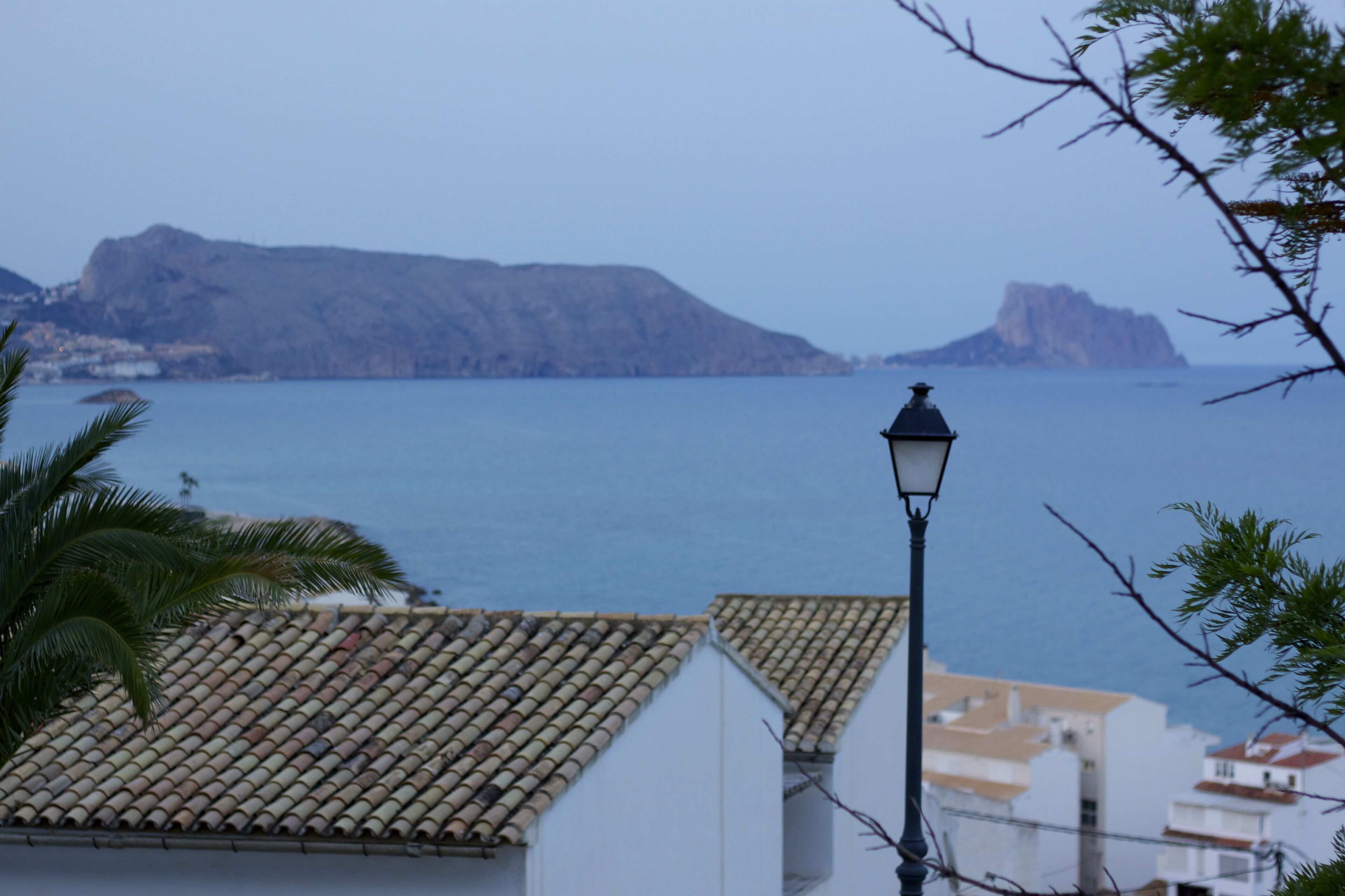 A coastal building with a tiled roof, a street lamp, and a view of the sea and distant mountains.
