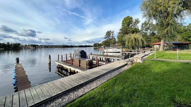A wooden dock extending into calm water, a boat docked at the pier, and a grassy area with trees and a building in the background.