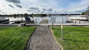 A wooden dock leading to a calm lake with boats, a grassy area, and a cloudy sky.