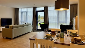 A modern living room with a sofa, dining table set for four, and large windows offering a view of a lake.