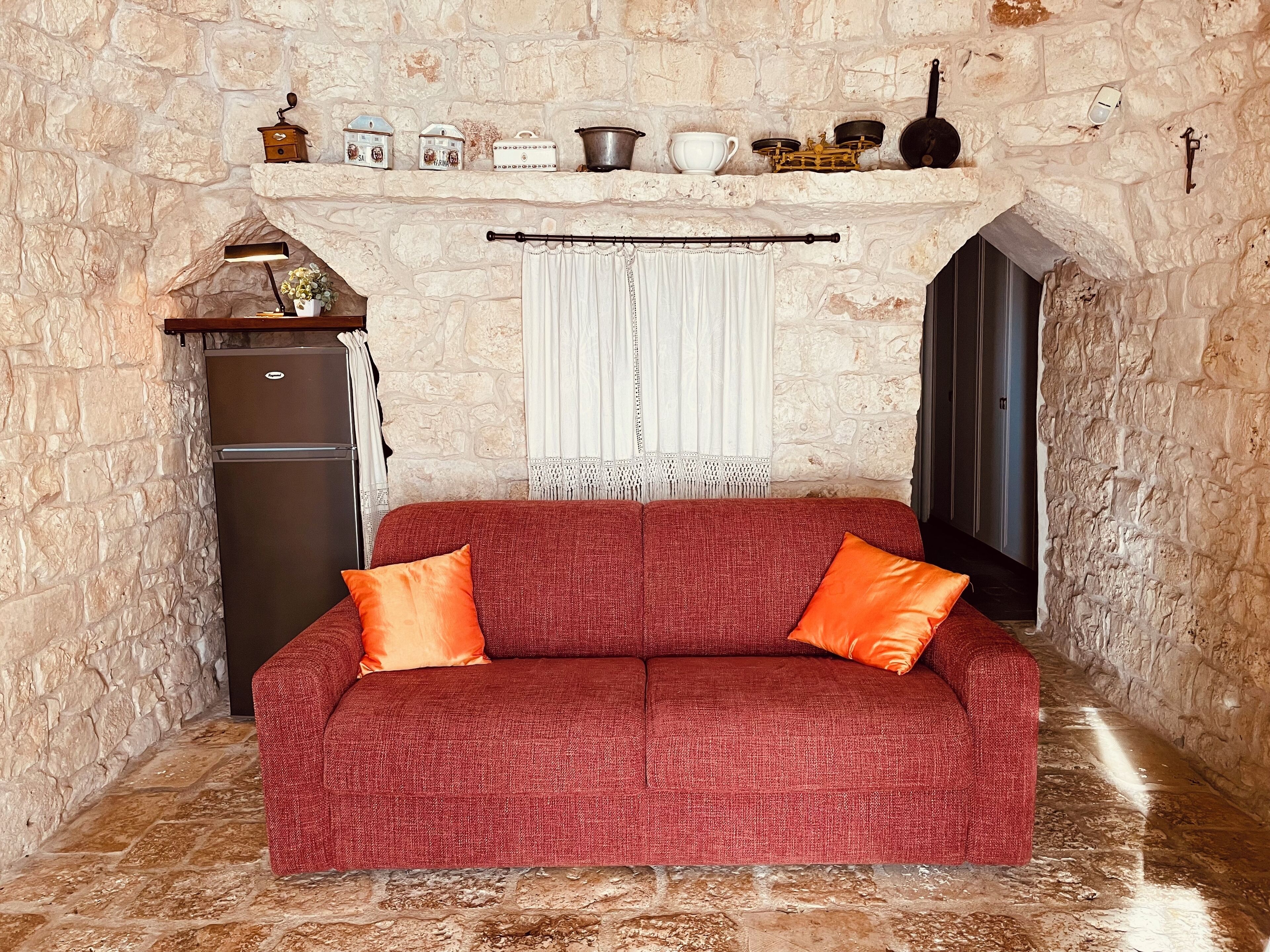 A cozy room with a red sofa, two orange cushions, a stone wall, a small shelf with kitchen items, a white curtain, and a door.