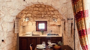 Un coin repas aux murs de pierre, avec une table en bois dressée pour deux personnes, une fenêtre au-dessus de l’évier et un rideau à motifs sur le côté droit.