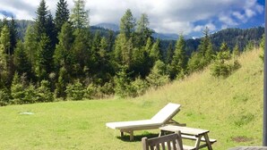 A wooden deck with a table and chairs, overlooking a grassy area with trees and a mountain backdrop.