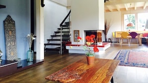 A spacious living room with a wooden floor, a central fireplace, a staircase, and a dining area with chairs.