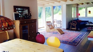 A spacious living room with a large wooden table, a sofa, and a colorful rug. There is a large window providing a view of the outdoors.
