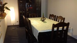 Ein Essbereich mit einem Tisch, gedeckt für sechs Personen, umgeben von Holzsesseln. Eine Lampe befindet sich links, und ein Bücherregal ist im Hintergrund zu sehen.
