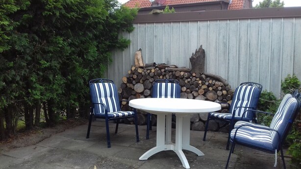 Eine Terrasse mit einem runden Tisch, vier Streifenstühlen, einer Steinmauer und einem Holzstoß.