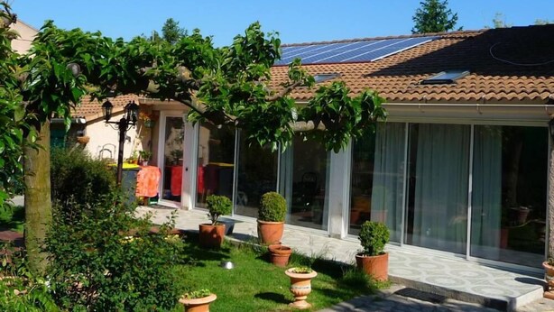 Une maison d’habitation avec un toit en tuiles, des panneaux solaires, un patio couvert et des plantes en pot.