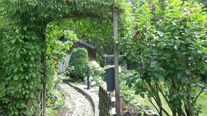 Un sentier de jardin mène à une maison en bois dotée d’un porche abrité, le tout entouré d’une végétation luxuriante et d’un mur de pierre.