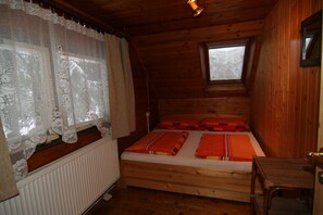 A wooden cabin room with a bed, a window with lace curtains, a small skylight, and a wooden chair.