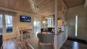 Tunnelmallinen huone, jossa on puinen katto, seinälle asennettu televisio, ruokapöytä tuoleineen, sohva ja pieni keittiöalue.