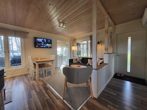Une pièce chaleureuse avec un plafond en bois, un téléviseur fixé au mur, une table à manger avec des chaises, un canapé et un petit coin cuisine.