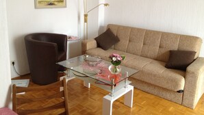 Ett vardagsrum med en beige soffa, ett soffbord i glas med en vas med blommor, en brun fåtölj och en golvlampa.