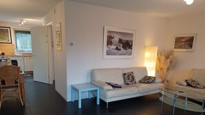 Uma sala de estar moderna com um sofá branco, uma mesa de centro de vidro e uma pequena mesa lateral. Há almofadas decorativas no sofá e um quadro emoldurado na parede.