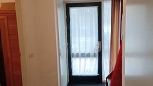 Um corredor com chão de azulejos escuros, uma porta à esquerda, uma janela com cortinas leves e uma cadeira vermelha perto da janela.