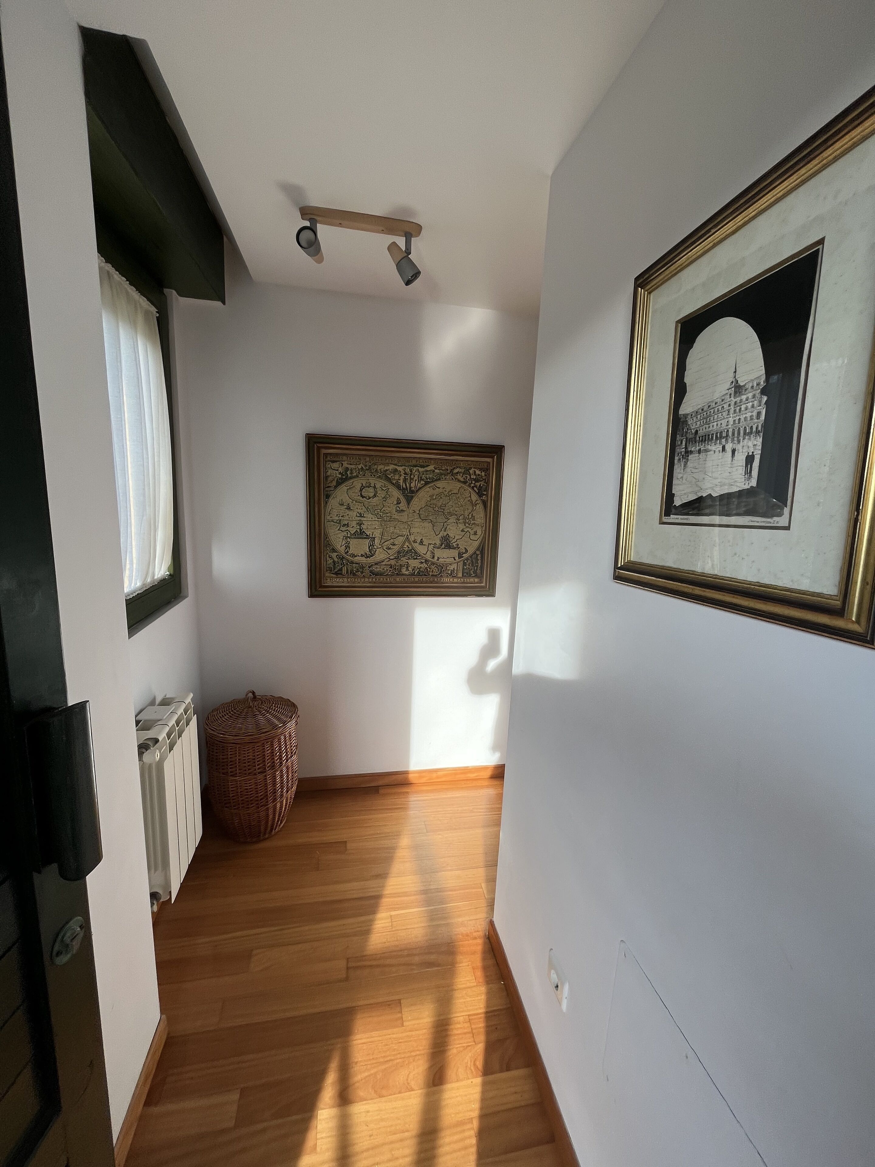 Un couloir avec un plancher en bois, un radiateur, un panier en osier et des œuvres d’art encadrées accrochées aux murs.