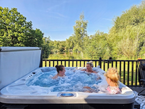Un jacuzzi con tres personas, rodeado por una terraza y vistas al lago.