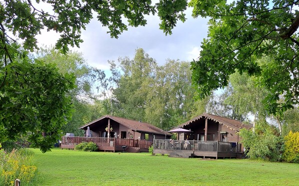 Two rustic wooden cabins with wooden decks and railings, surrounded by lush green grass and trees. - Lakeside Lodge with 6 berth hot tub and private fishing peg (Tattershall)