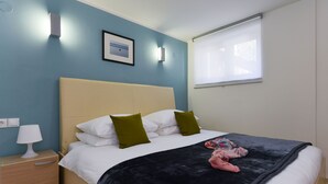 A neatly made bed with a black blanket, two white pillows, and a green pillow. There is a bedside table with a lamp and a power outlet. A framed picture hangs on the wall above the bed. - Apartment Mare with beautiful sea views (Lun)
