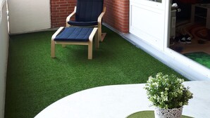 A balcony with artificial grass, a wooden chair, a small table with a potted plant, and a sliding glass door leading to an indoor area.