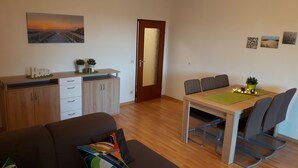 A modern living room with a sofa, a dining table with chairs, a kitchen area with cabinets, and a door.