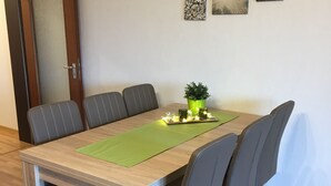 A dining area with a wooden table, six chairs, a green placemat, a potted plant, and two wall-mounted artworks.