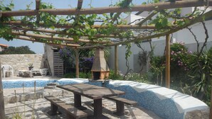 Un patio de piedra con una mesa y bancos de madera, rodeado de un muro de piedra y una pérgola de madera cubierta de plantas trepadoras.