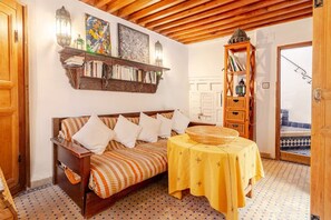 Une pièce chaleureuse avec un plafond en bois, un lit recouvert d’une courtepointe à carreaux, une bibliothèque, une petite table recouverte d’une nappe jaune et un sol à motifs.