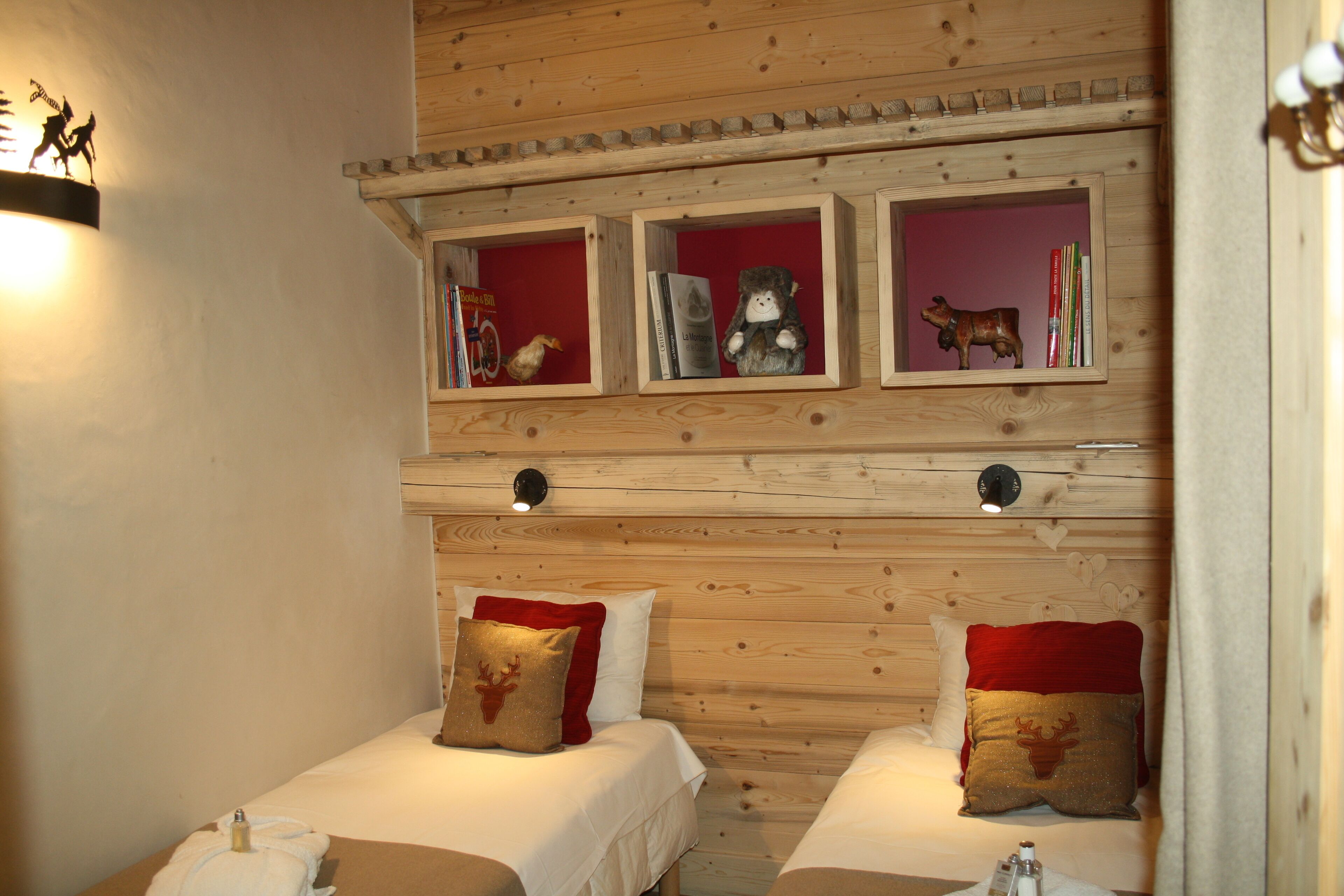 A cozy room with two beds, wooden headboards, and shelves above containing books and decorative items.