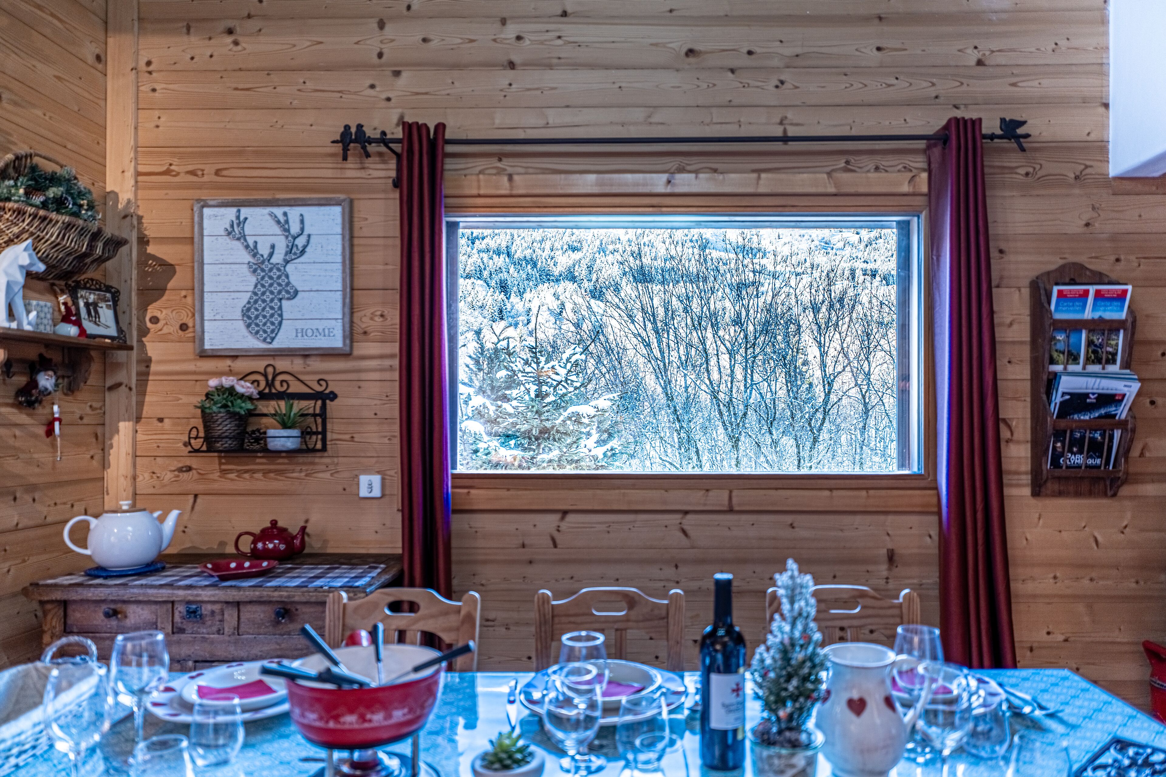 A wooden cabin interior with a dining table set for four, a large window with a snowy view, and various decorations including a framed deer head and a basket on the wall.
