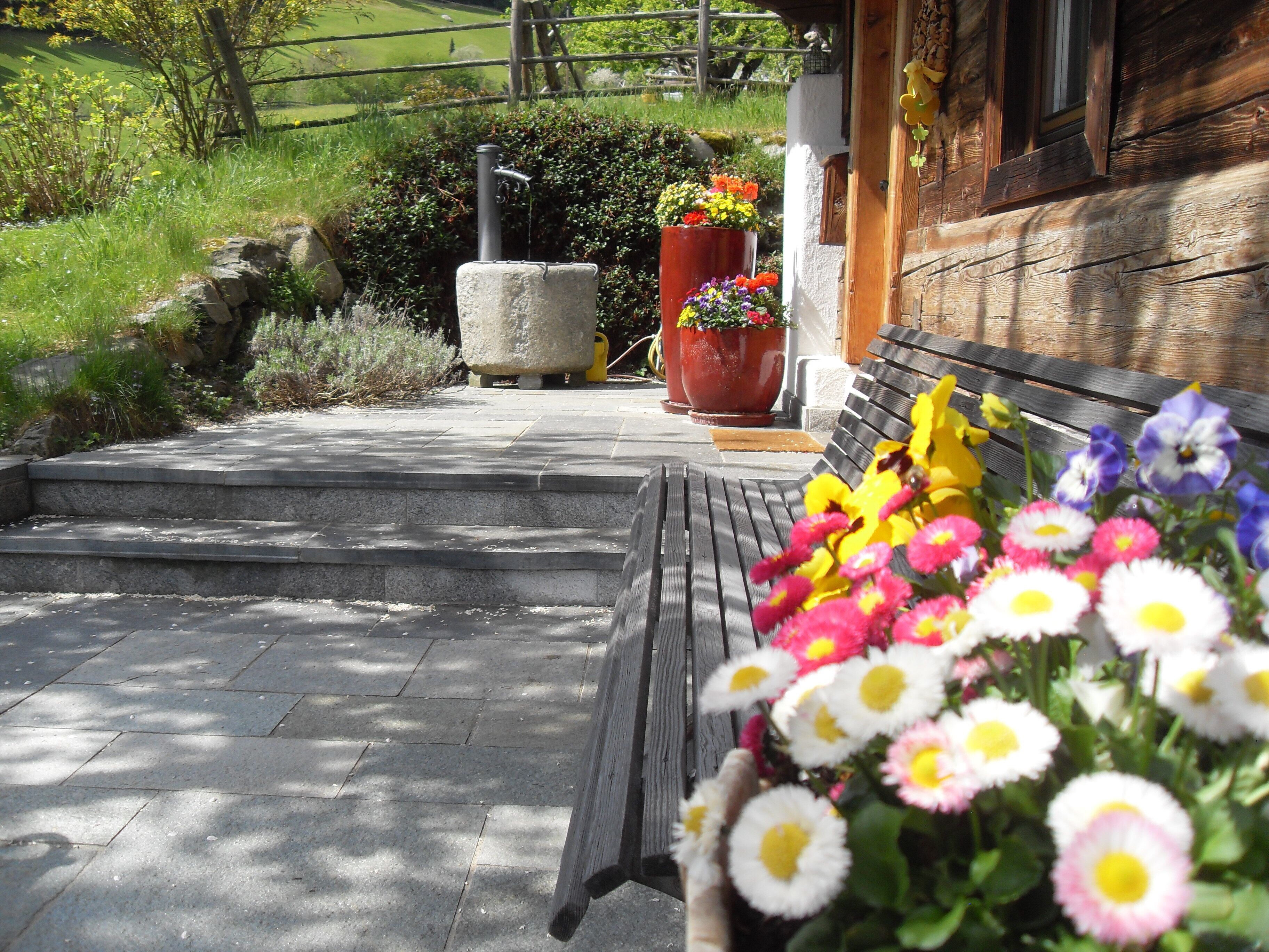 Un banc en bois avec un panier de fleurs devant, des marches en pierre menant à un bâtiment et des plantes en pot.