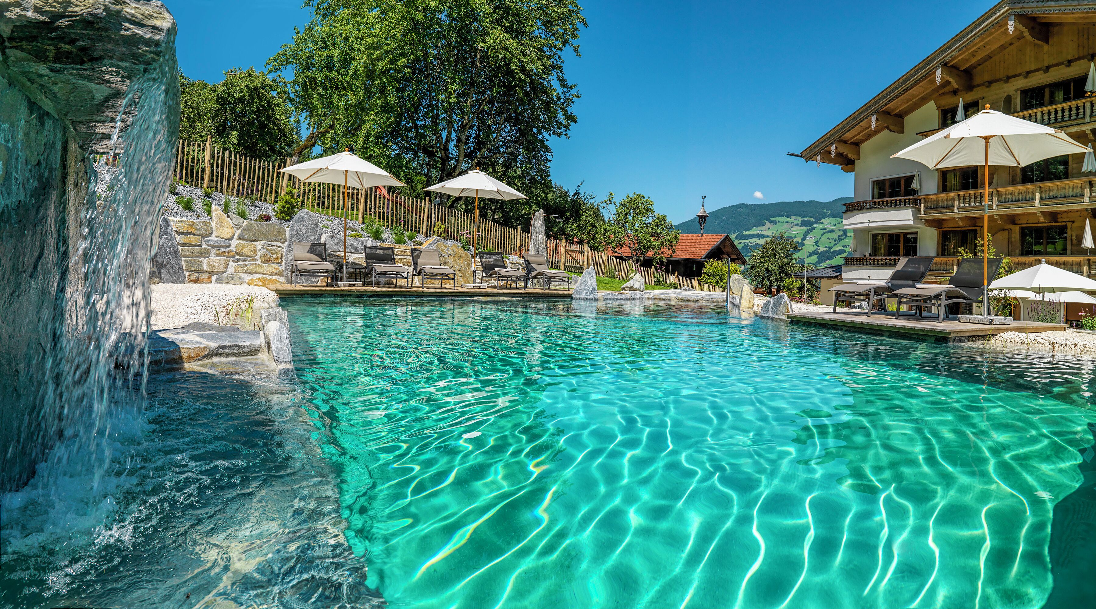 Une piscine aux eaux cristallines, bordée de chaises longues et d’ombrelles, et entourée de murs de pierre et d’une clôture en bois.