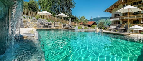 A clear pool with lounge chairs and umbrellas, surrounded by stone walls and a wooden fence.