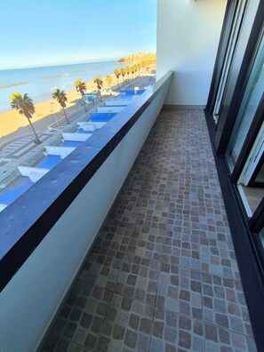 Ein Balkon mit Fliesenboden, Blick auf einen Strand mit Palmen und ein Poolbereich.