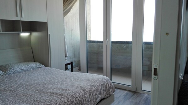 A bedroom with a bed, a chair, a sliding glass door to a balcony, and a ceiling light.