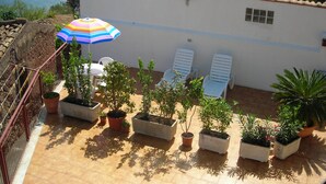 Eine Terrasse mit Topfpflanzen, einem gestreiften Sonnenschirm und zwei weißen Liegestühlen.