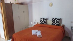 Ein ordentlich gemachtes Bett mit orangefarbener Bettdecke und zwei dekorativen Kissen, ein Holzschrank und ein kleiner Tisch mit Lampe.