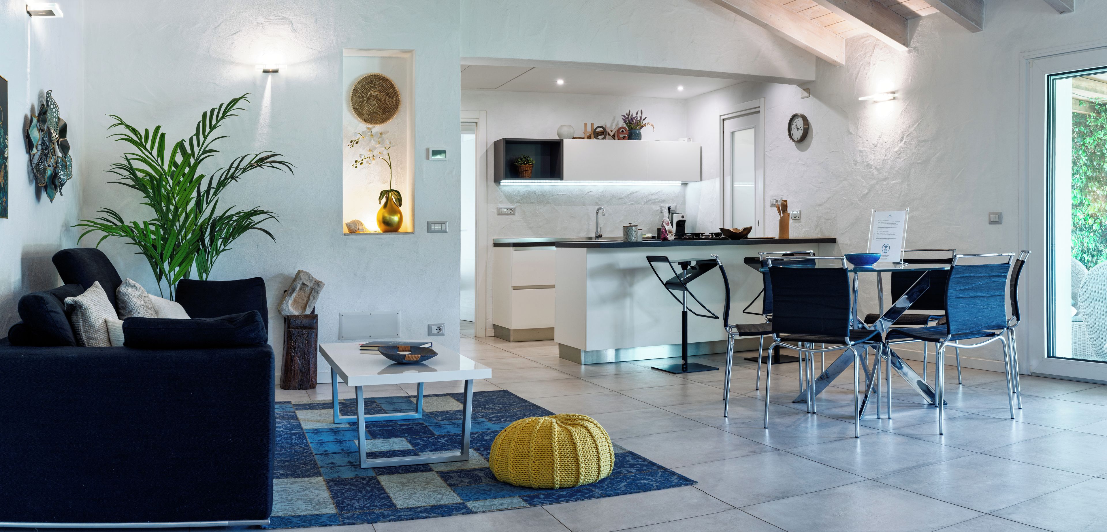 Un salon moderne comprenant un canapé, une table basse et des fauteuils, un coin cuisine avec un bar, et une grande fenêtre laissant entrer la lumière naturelle.