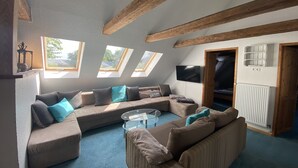 A cozy living room with a sectional sofa, a glass coffee table, and a TV mounted on the wall. There are skylights allowing natural light to enter. - North Sea vacation right before SPO; 6-8 people, large garden apartment skipper - (Tating)