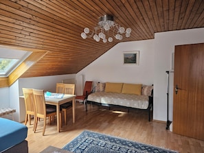 Ein gemütliches Dachzimmer mit Holzbalkendecke, einem Esstisch mit Stühlen, einer Couch und einer Pendelleuchte.