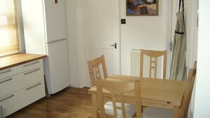 A compact kitchen with wooden cabinets, a white refrigerator, a dining table with chairs, and a painting on the wall. - Beautiful 3 bedroom apartment in London (Tooting)