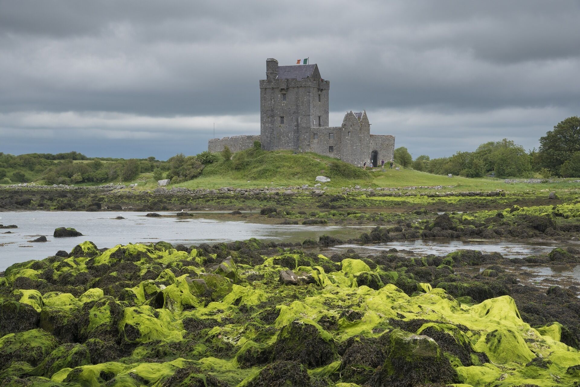A historic stone castle on a grassy hill with a rocky shoreline in the foreground.