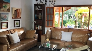 Een woonkamer met een bruine leren bank, een glazen salontafel en een zwart-wit fauteuil. Er is een boekenkast met boeken en decoratieve objecten, een kroonluchter en een raam met uitzicht op groen.