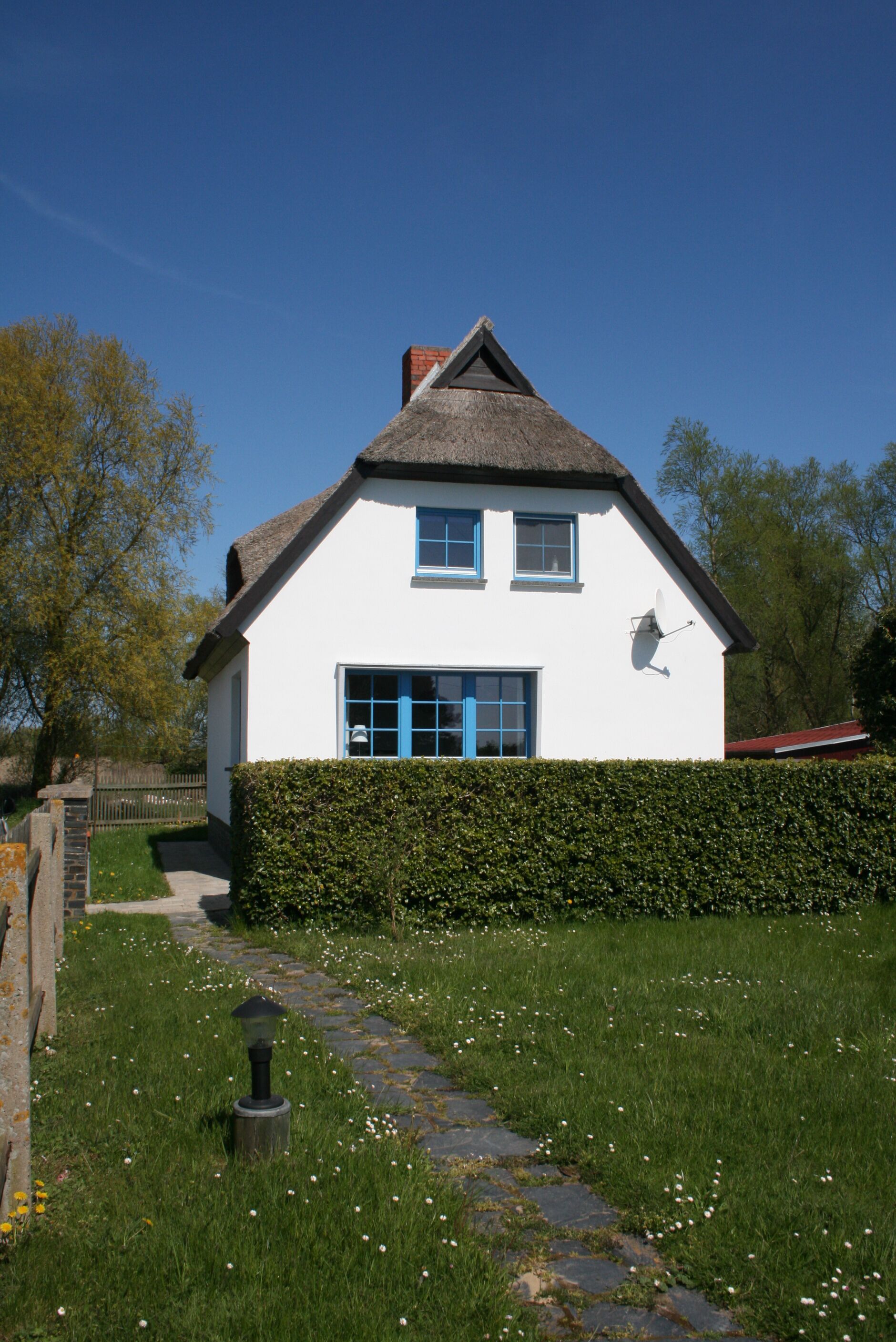 Une maison blanche avec un toit de chaume, une haie devant et un chemin de pierres menant à l’entrée.
