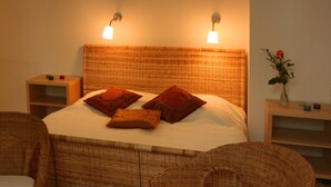 A neatly arranged bedroom with a large bed, two bedside tables, a nightstand, a chair, a small table with a candle, and a vase with a single flower.