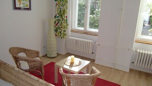 A room with a bed, wicker chairs, a table with a candle, a radiator, and a window with a view of greenery.