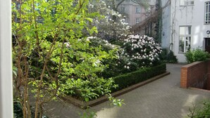 A paved pathway flanked by lush greenery and flowering bushes, leading to a building with a brick facade and multiple windows.